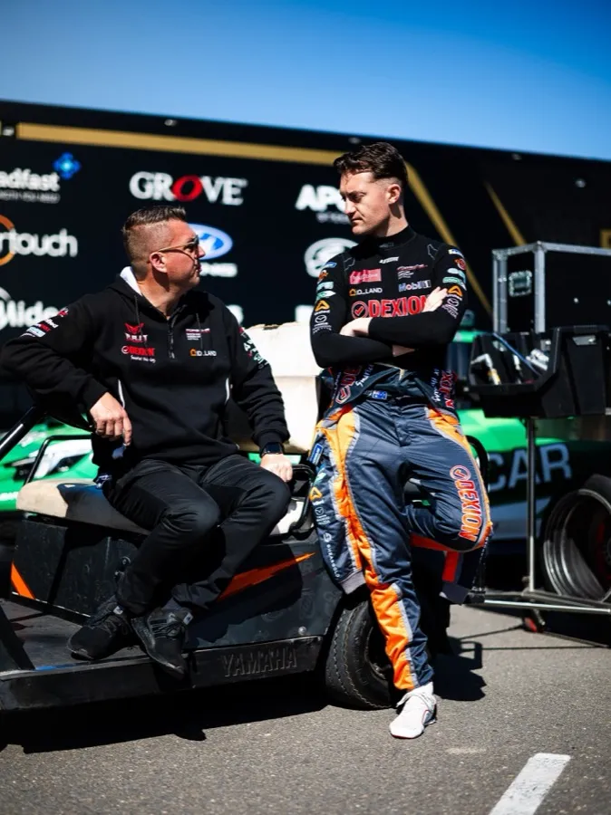 A driver in a race suit and a team member leaning on equipment in the pit lane