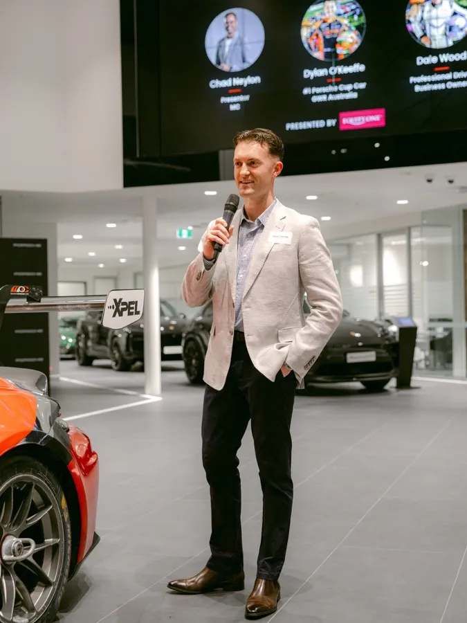 Dylan O’Keeffe speaking into a microphone at an Equity-One event beside a Porsche